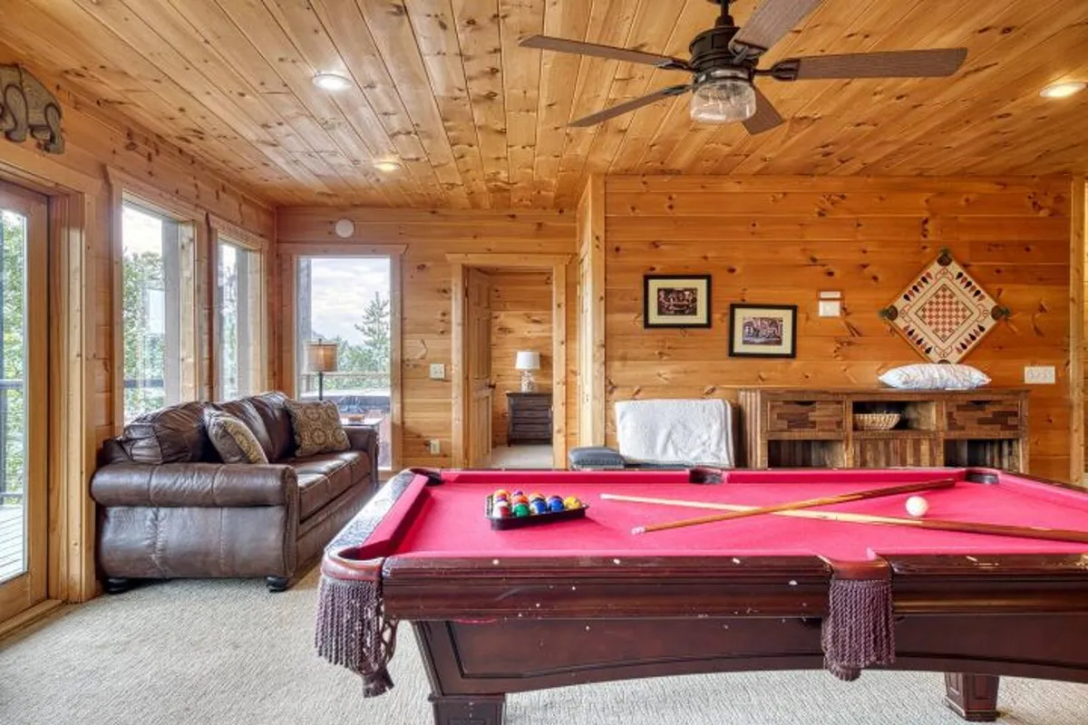A well-appointed game room featuring a pool table at the center, complemented by a leather couch along the wall and windows that let in natural light. The warm wooden walls create an inviting environment for friendly competition and socializing.