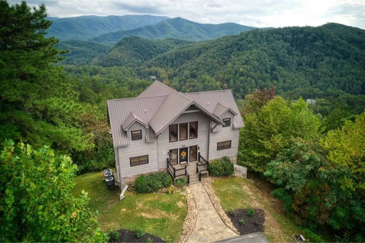 This inviting exterior image shows the estate nestled in the Smoky Mountains, surrounded by lush trees and landscaped paths. The house is set against a backdrop of rolling mountains, hinting at the proximity of hiking trails and panoramic views. The scene conveys an atmosphere perfect for family gatherings, outdoor barbecues, and enjoying the tranquility of nature.