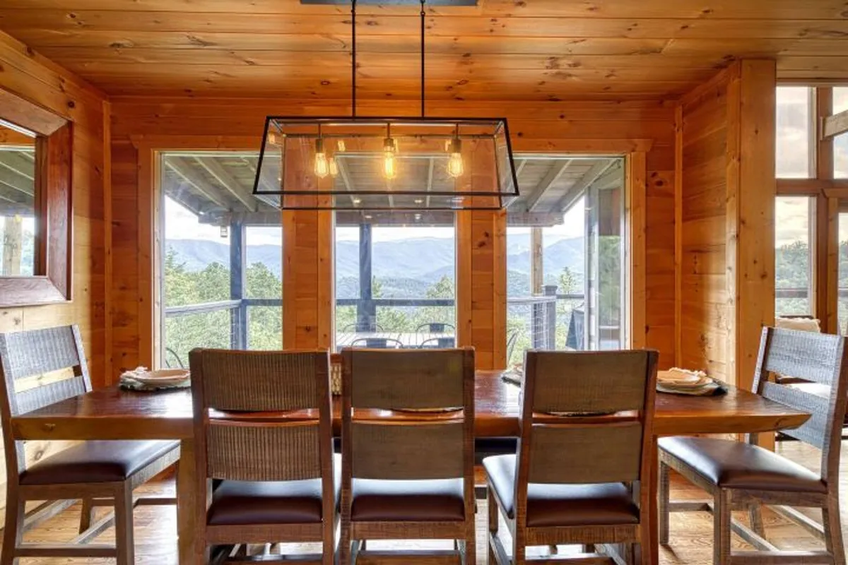 A bright, welcoming dining area with a large wooden table, stylish hanging light fixture, and panoramic windows opening to breathtaking mountain views. The setting is perfect for group meals and shared moments.