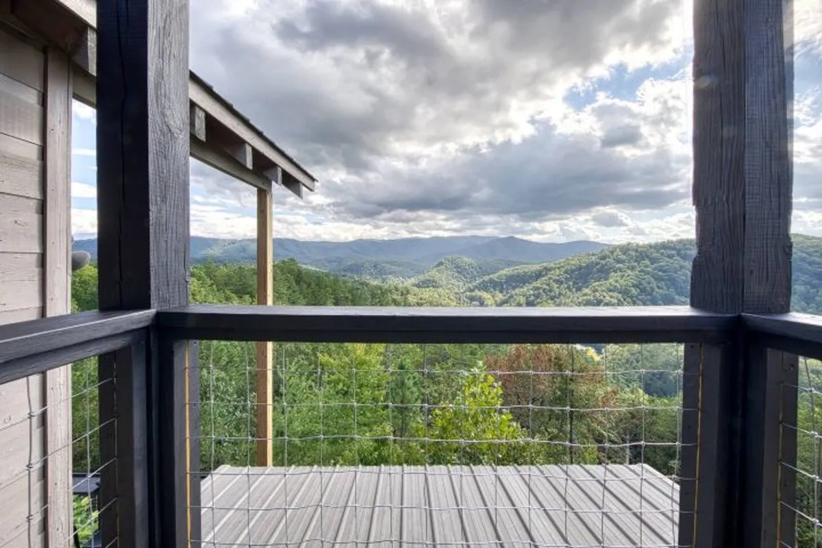 A scenic deck view overlooking rolling, forested mountains beneath a cloudy sky. The image captures a wooden deck railing in the foreground, inviting guests to step outside and immerse themselves in nature. The serene atmosphere is accentuated by lush green trees and the distant peaks, suggesting a perfect spot for morning coffee or evening stargazing.