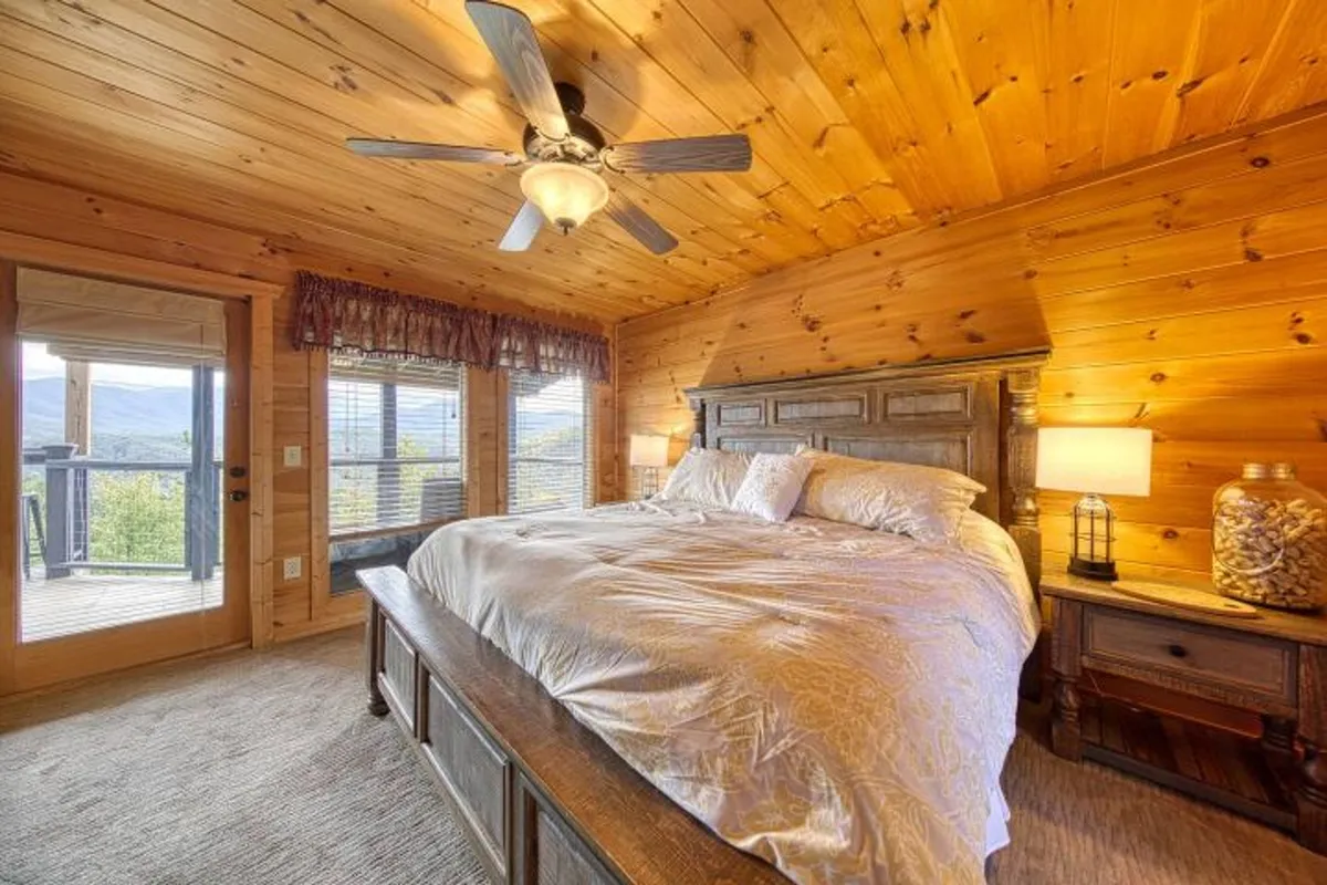 A spacious, tranquil bedroom featuring a king-sized bed with plush linens, an elegant wooden ceiling, and large windows that offer breathtaking mountain views. Soft lighting from a nightstand lamp adds to the serene, restful atmosphere, while hints of a private balcony outside suggest a seamless connection to nature.