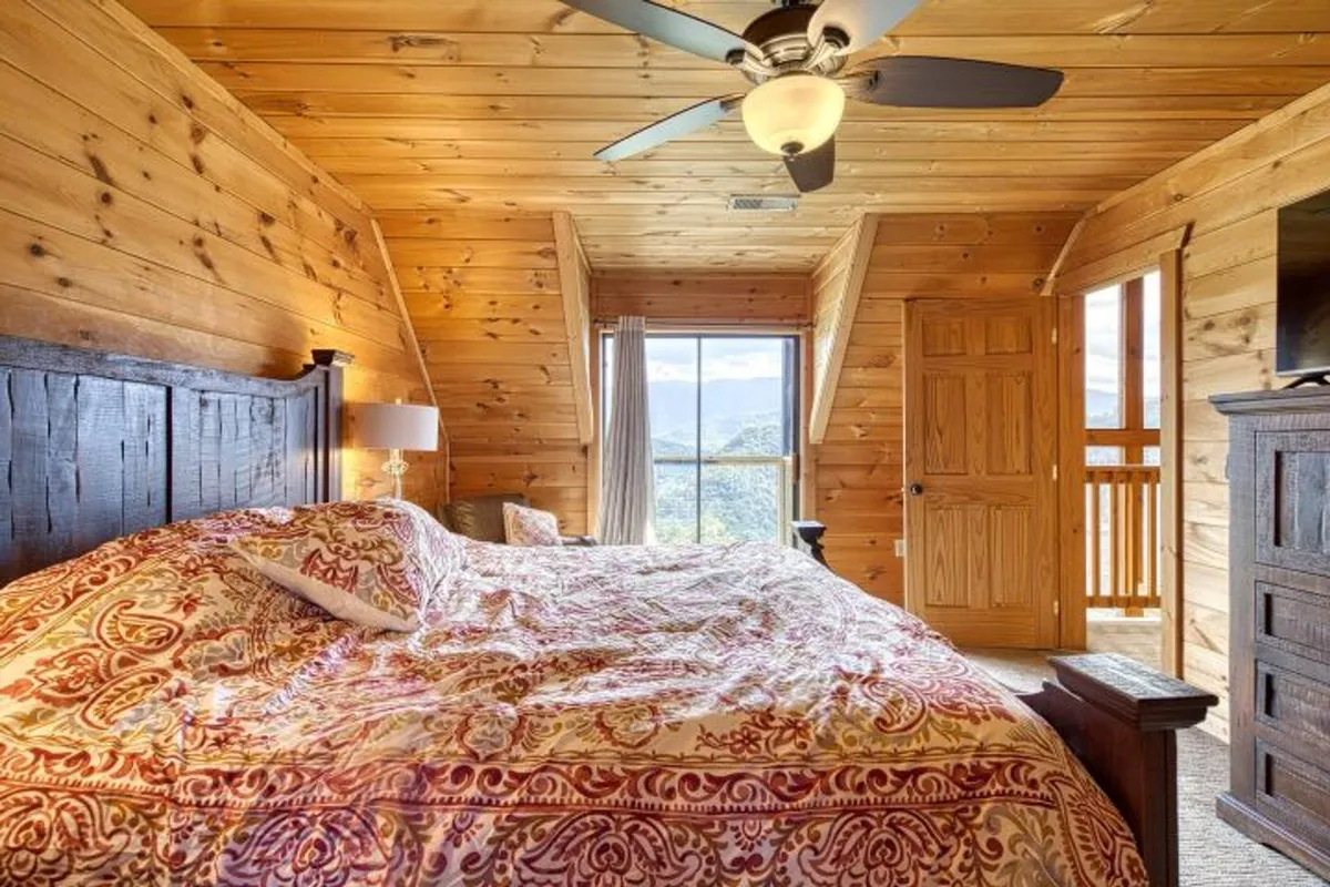 A serene bedroom highlighted by a comfortable bed with ornate bedding positioned next to a large window framing sweeping mountain views. The wooden ceiling and walls enhance the cozy, natural ambiance, and a bedside lamp invites quiet evenings and slow mornings.