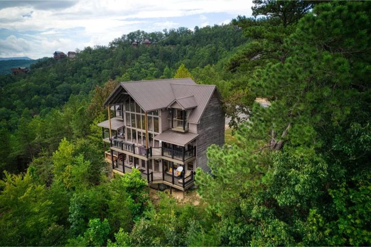 A modern wooden cabin exterior with expansive glass windows is nestled in verdant woods. The porch and surrounding greenery invite outdoor relaxation, and the design exudes sophistication and modern elegance. The space is perfect for sunset viewing or peaceful mornings immersed in nature.