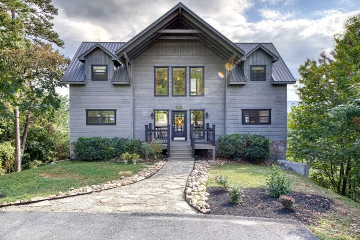 The image showcases the grand front facade of a luxurious cabin nestled in lush greenery, with impressive wall-to-ceiling windows offering panoramic mountain views, a charming stone pathway, and a spacious porch perfect for watching sunsets or hosting gatherings.