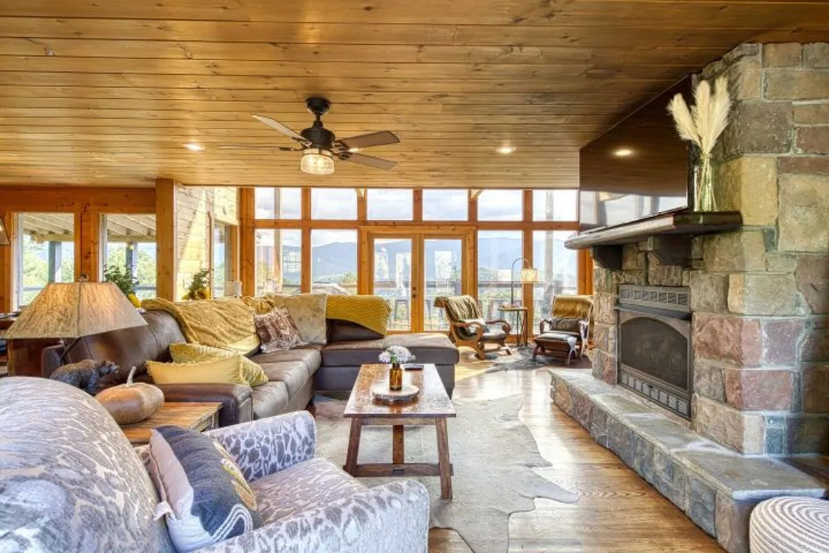 A warm, inviting living room with a large sectional sofa, stone fireplace, and expansive windows framing magnificent mountain views. The space features a wooden coffee table and tasteful decor, offering plenty of room for socializing and relaxation.