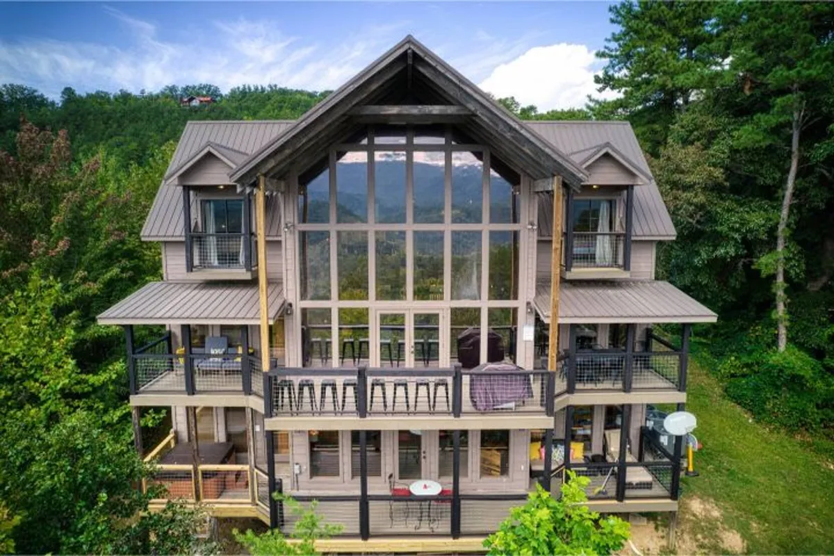 The image features the exterior of a luxurious cabin, set amidst lush greenery and framed by large glass windows and balconies. The sunset light glows on the house, suggesting warmth and exclusivity. The inviting balconies and outdoor lounge areas overlook sweeping natural vistas, creating an atmosphere of indulgence and tranquility.