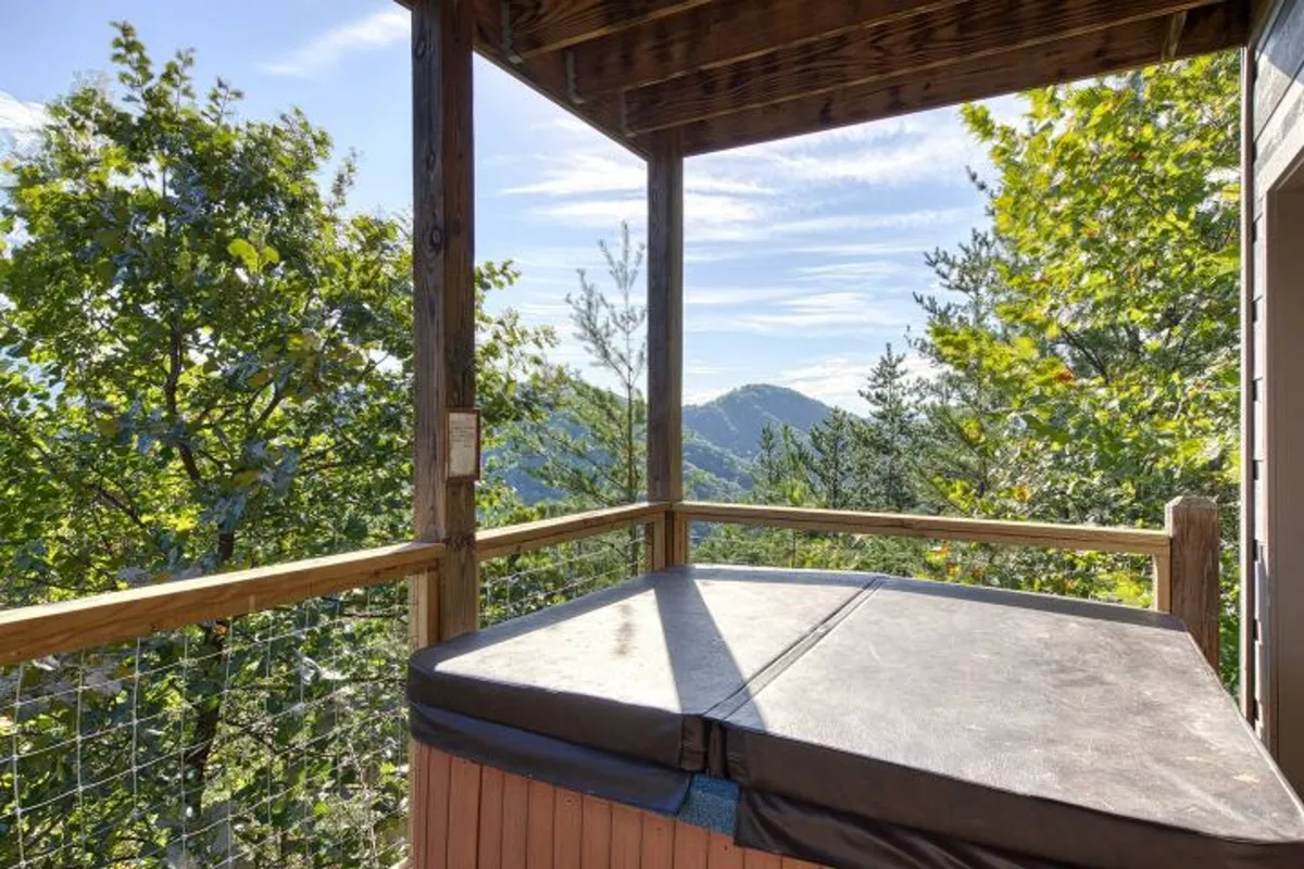 A peaceful outdoor deck featuring a bubbling hot tub with a direct view of the rolling Smoky Mountains. The wooden railing adds rustic charm, and the surrounding greenery offers privacy and a seamless connection to nature. The hot tub is positioned for guests to enjoy both sunrise and sunset mountain vistas.
